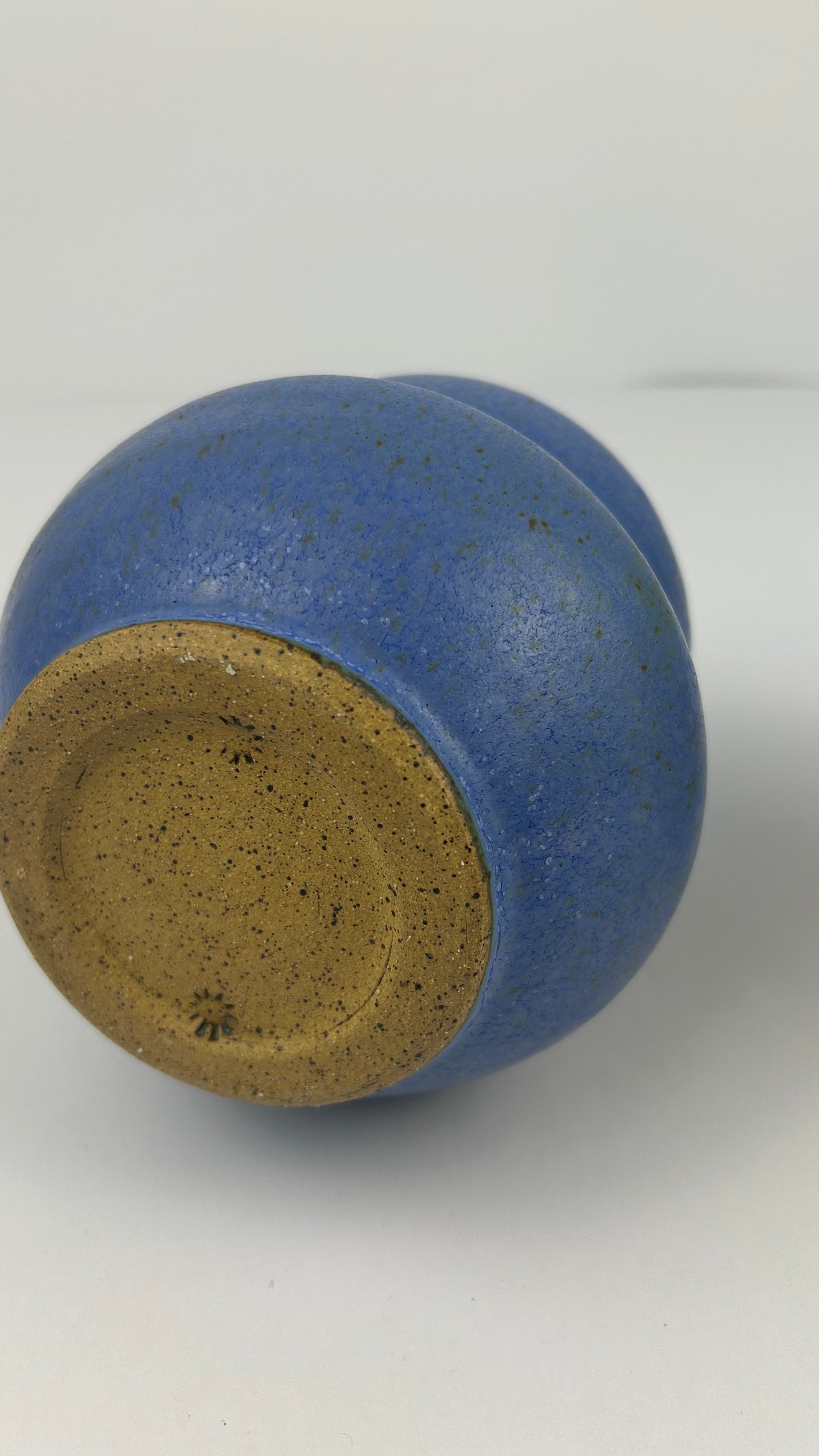 A close-up of a ceramic vessel's bottom, showing a speckled blue glaze over a natural tan clay base, photographed on a light gray background.
