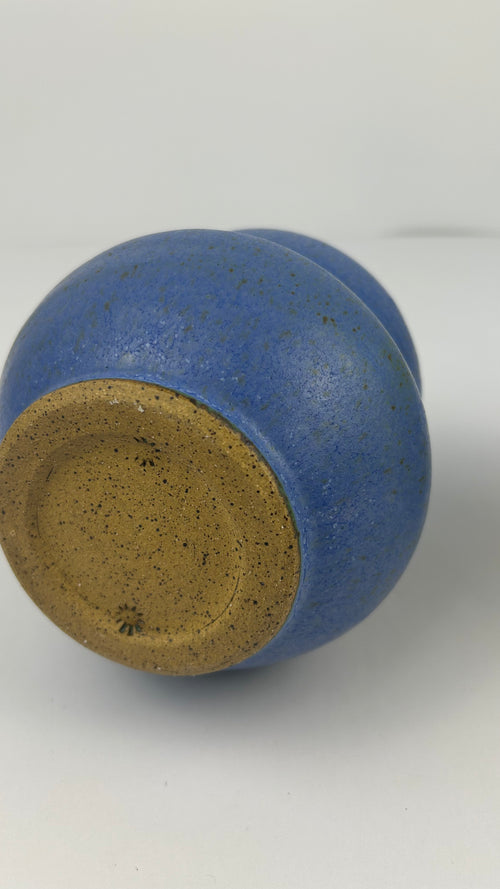 A close-up of a ceramic vessel's bottom, showing a speckled blue glaze over a natural tan clay base, photographed on a light gray background.