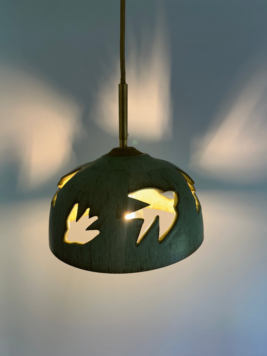 Stoneware green speckled pendant lamp with bird piercings and gold cord with shadows on the wall