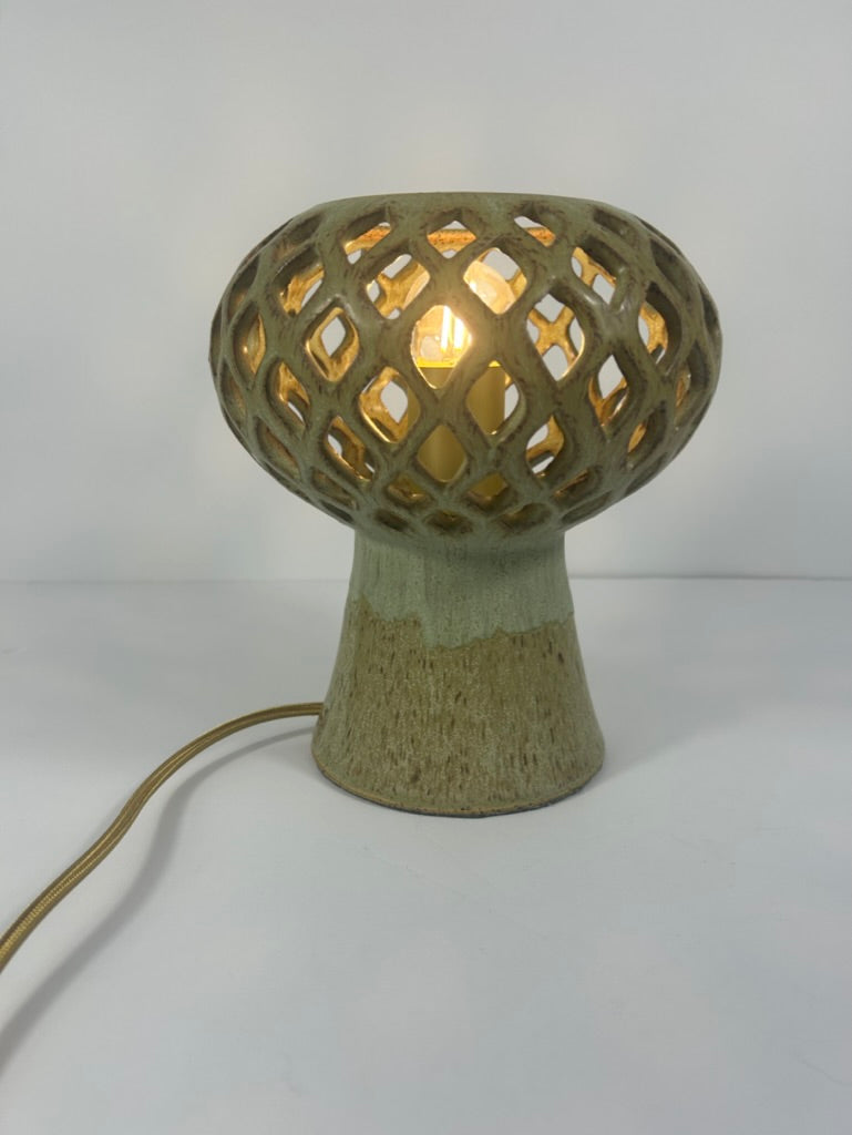 A ceramic table lamp with a textured, earthy green and beige base and a lattice-patterned ceramic shade that glows warm yellow from an internal light bulb. A gold-toned power cord extends to the left on a plain white background.