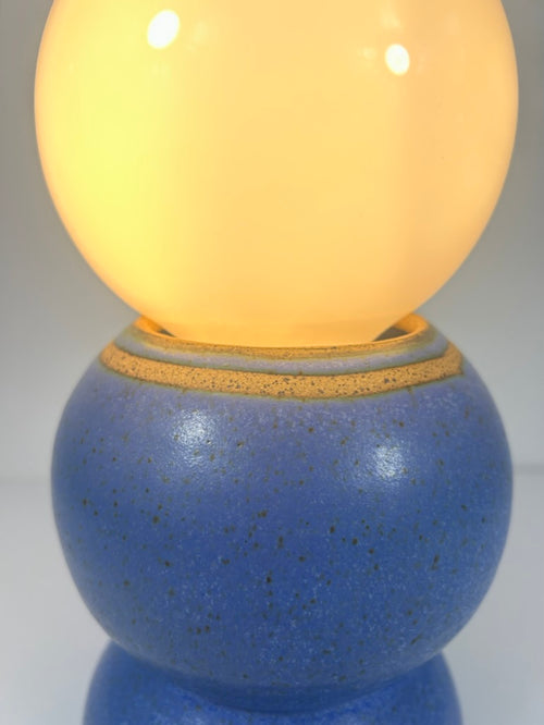 A close-up of a decorative table lamp with a glowing warm yellow globe on top of a speckled blue ceramic base with a gold band detail.
