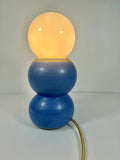 A modern ceramic bubble table lamp with a glowing warm yellow-orange spherical top, a stacked blue ceramic base, and a gold-toned cord, photographed against a plain light gray background.