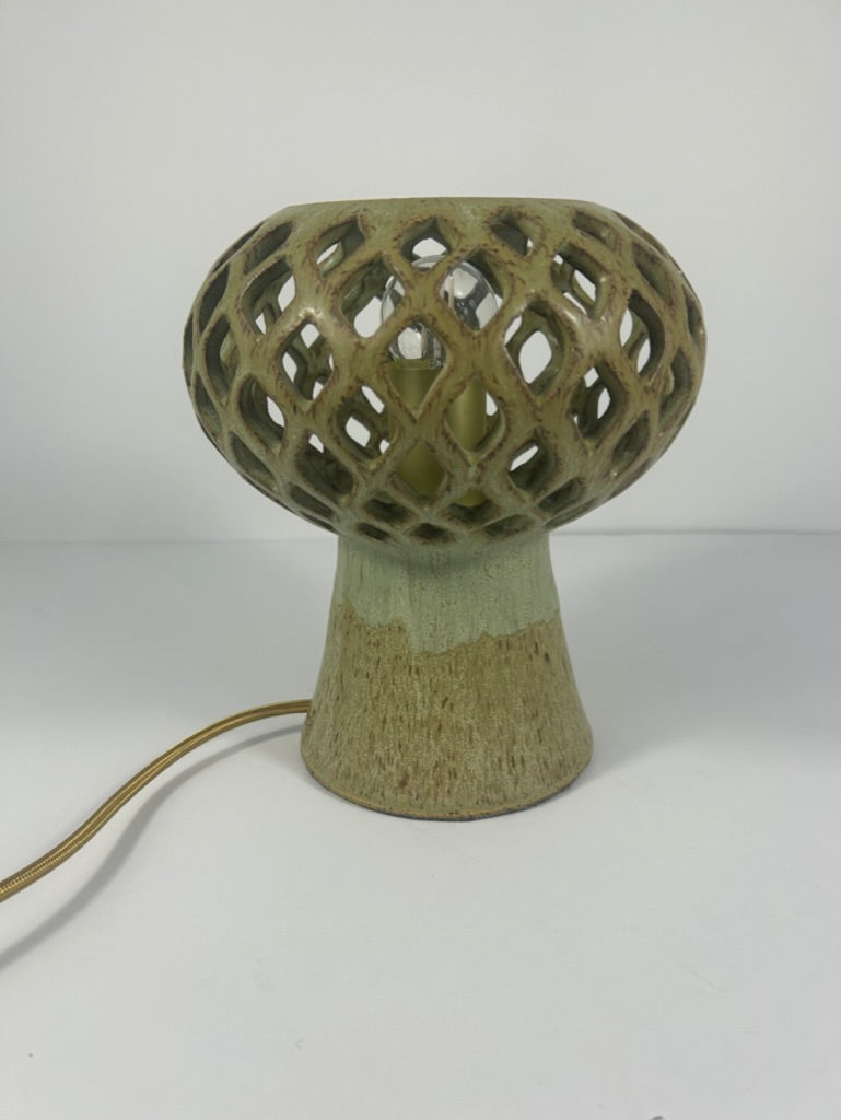 A ceramic table lamp with a textured, earthy green and beige glaze. The lamp features a wide, ceramic pierced geometric shade on top of a tapered base, with a gold-toned power cord extending to the left, set against a plain white background
