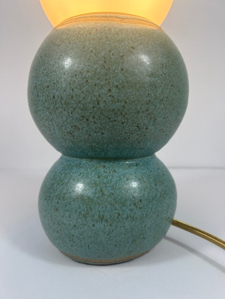 A two-tiered ceramic table lamp with a speckled teal-green glazed finish, emitting a warm yellow light from the top, shown against a plain white background.