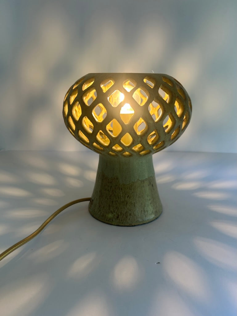 A mushroom-shaped table lamp with a pierced ceramic shade and a textured green base, glowing with warm light on a white surface that casts leaf-like shadows.
