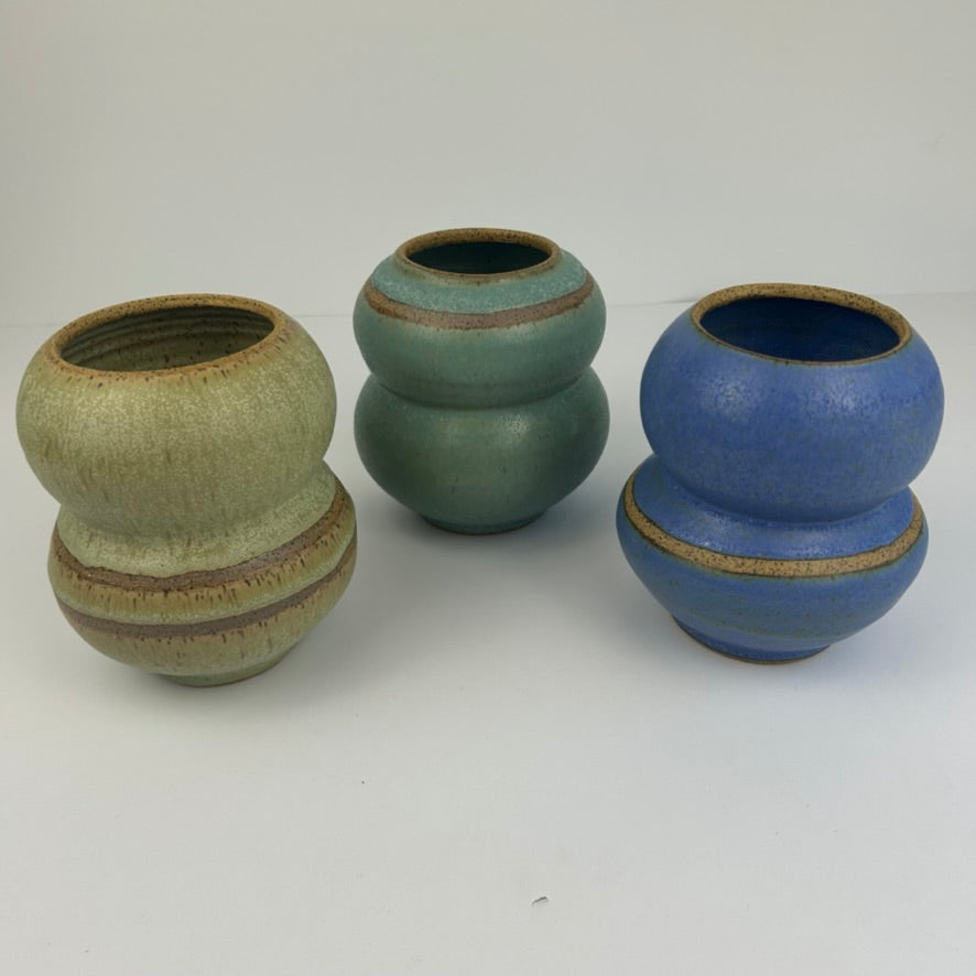 Three stacked ceramic vases with rounded, gourd-like forms on a plain white studio background. The left vase has a muted sage-green glaze with brown edging, the center vase is teal-green with brown edging, and the right vase is medium blue with brown edging.