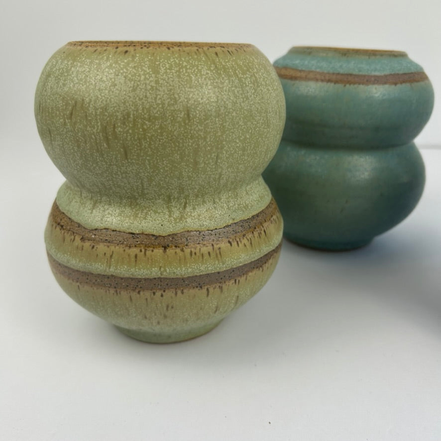 Two handmade ceramic vases with a textured glaze, one in light olive green and the other in teal blue-green, both featuring earthy brown bands, set against a plain white background.