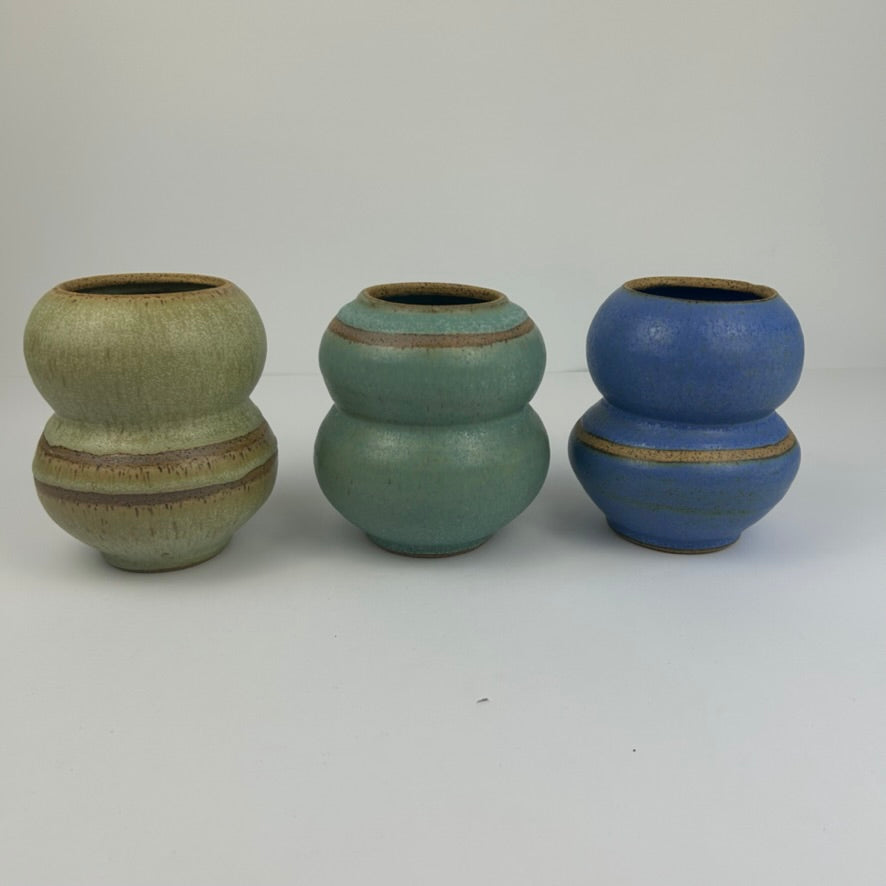 Three handmade ceramic double-bulb vases with stacked rounded forms, displayed side-by-side on a light gray background. The vase on the left has a textured sage green glaze with brown earthy bands, the middle vase has a muted teal-green glaze with brown bands, and the vase on the right has a vibrant cobalt blue glaze with a thin gold-toned band.