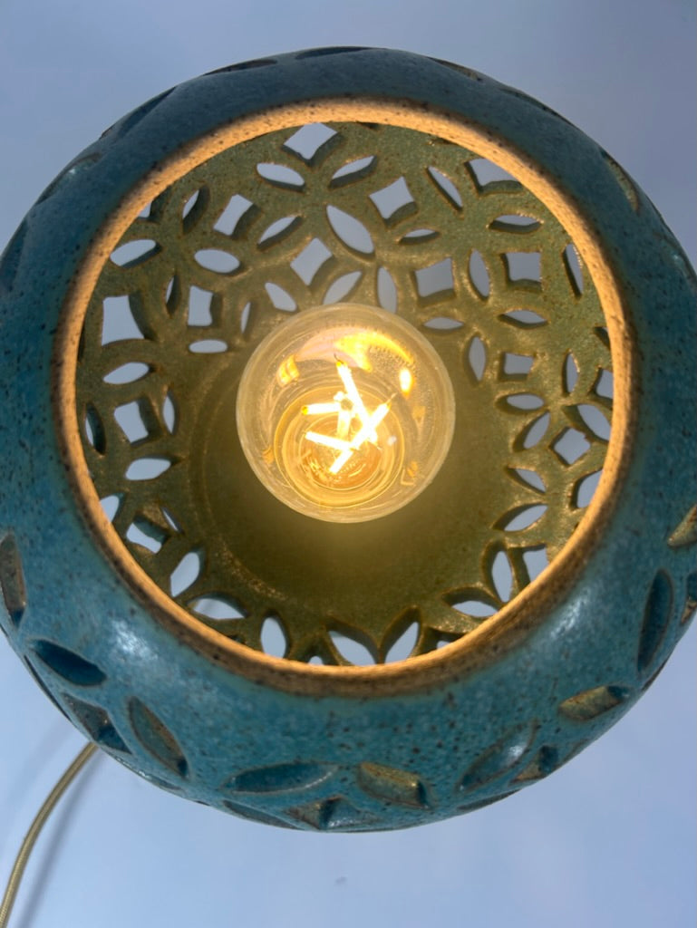Close-up of a teal ceramic lamp shade with a glowing warm light bulb inside, showing a cutout geometric pattern.