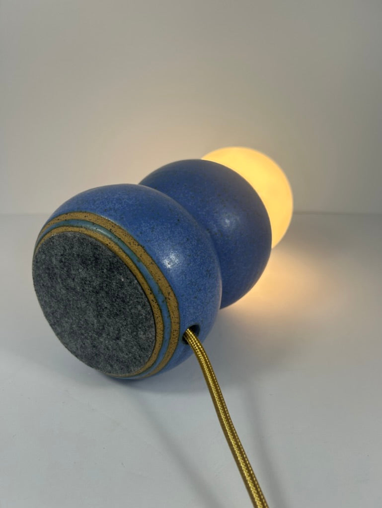 A blue ceramic table lamp with a spherical body, a glowing warm white bulb, a textured gray circular end, and a gold cord, photographed on a neutral light gray background.