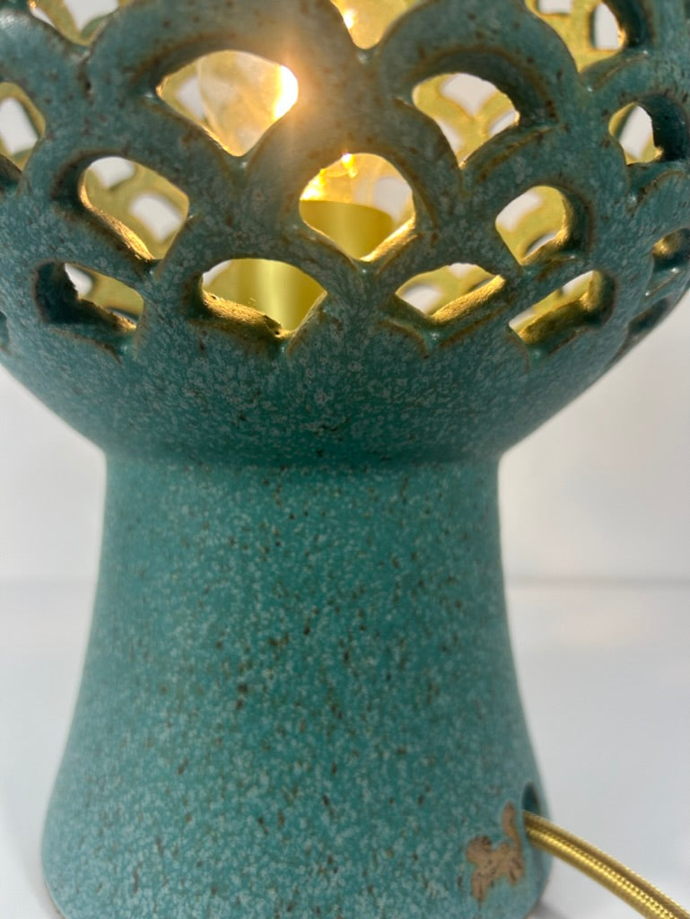 Close-up of a teal ceramic lamp with a textured speckled finish, featuring a scalloped cutout top and a warm yellow light glowing inside, with a gold-toned cord visible at the base.