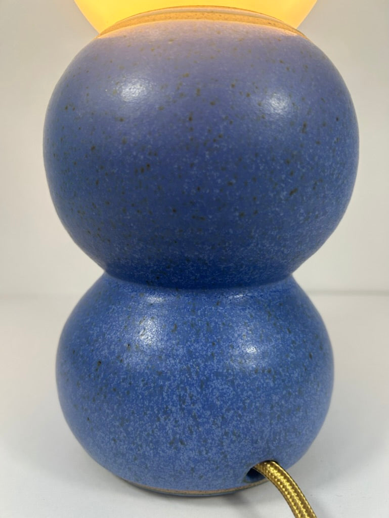 A blue speckled ceramic table lamp with a double-bulb design, a glowing yellow top shade, and a gold cord, shown against a plain light background.