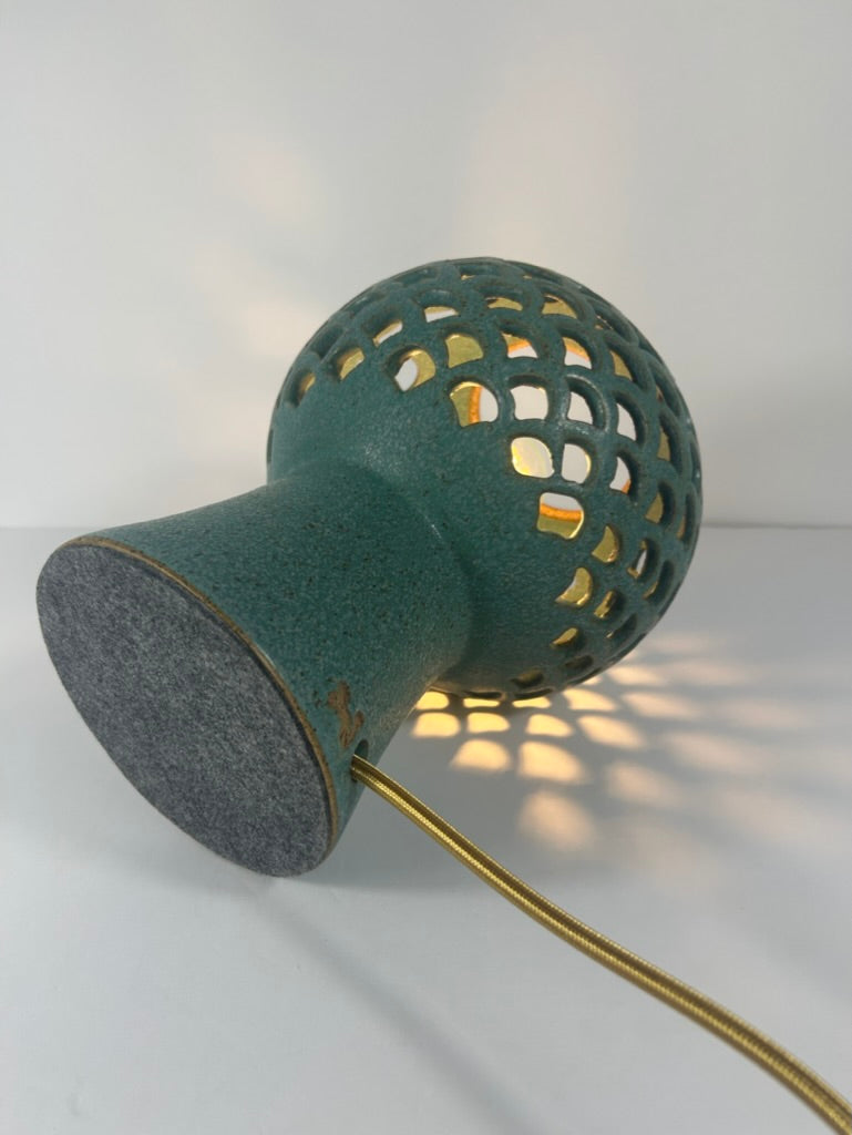 A modern sculptural table lamp with a textured teal-green ceramic body, featuring a pierced spherical ceramic shade and a gray felt-like base, with a gold-toned cord, shown on a white background.