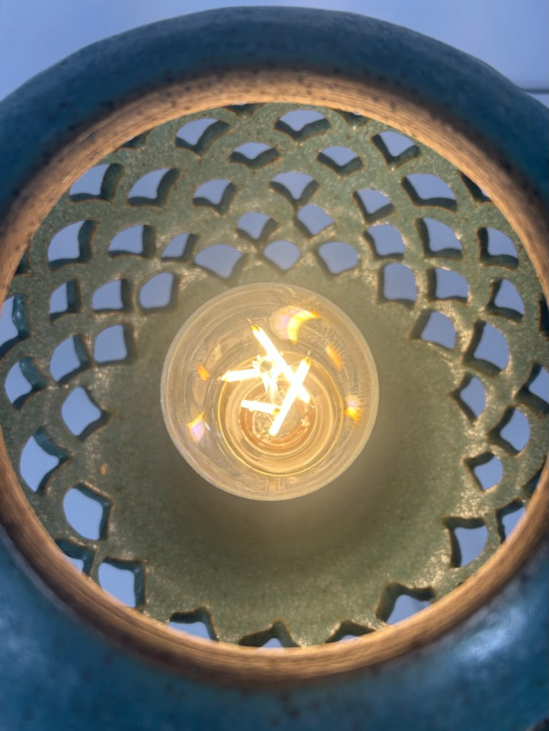 Close-up of a decorative ceramic lamp shade with a glowing light bulb visible through a circular cutout pattern.