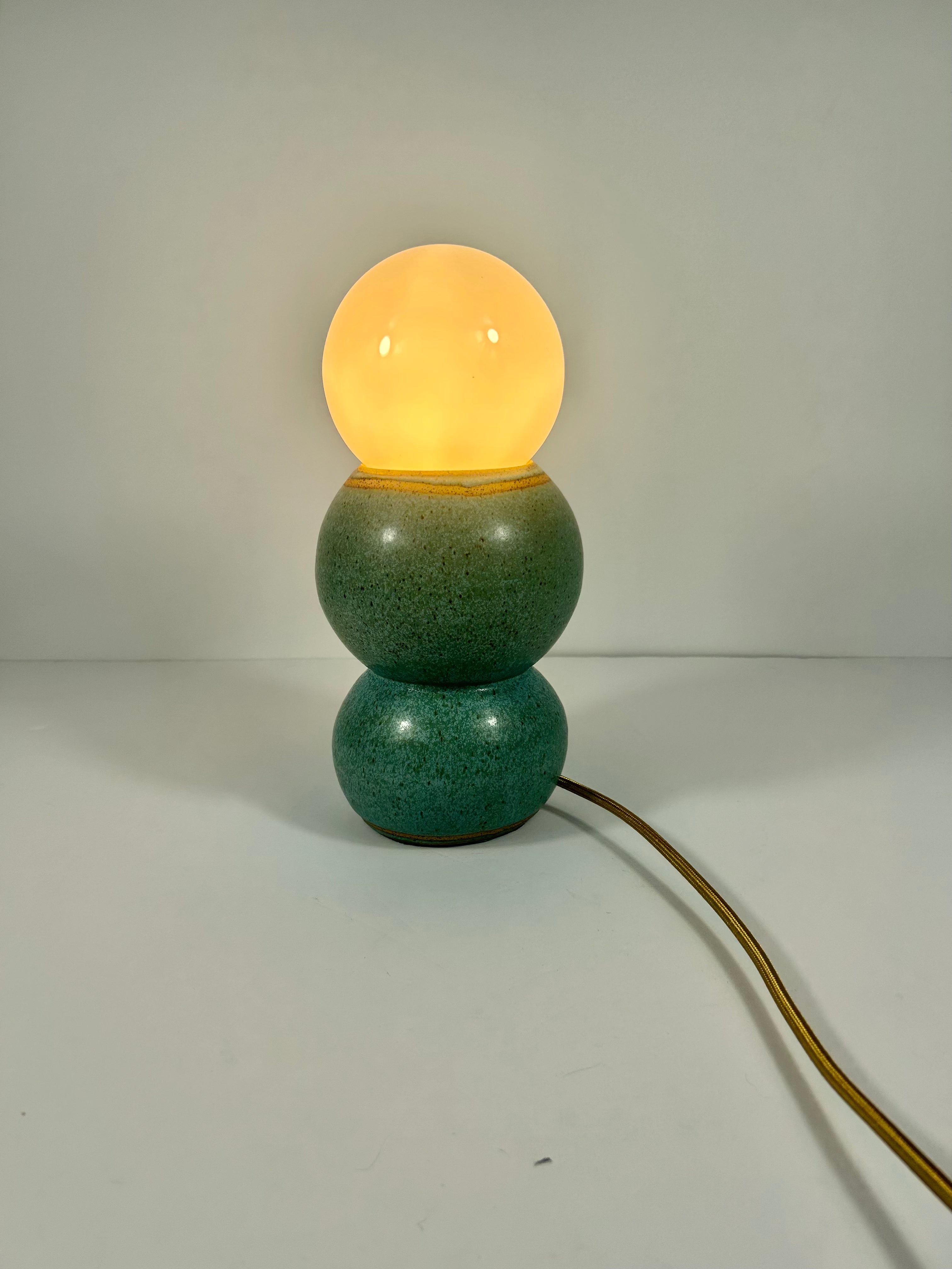 A vintage-style ceramic table lamp with a two-tiered blue-green speckled ceramic base and a glowing warm yellow vintage globe shade, shown on a plain light gray background with its power cord visible.