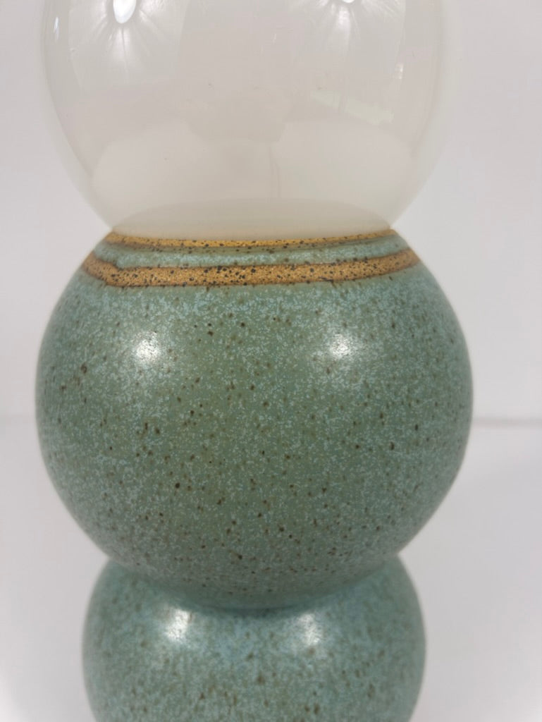 A close-up of a decorative ceramic table lamp with a speckled blue-green green body, a warm tan band near the top, and a frosted white rounded upper section.