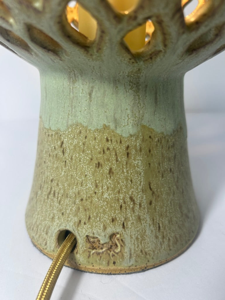 Close-up of a ceramic table lamp base with a textured, speckled glaze in pale green and beige, featuring a gold-toned cord plug and warm light glowing through the openwork top.