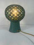 A teal ceramic table lamp with a pierced spherical shade and a flared base, glowing with a warm light from within. A gold-toned power cord extends from the side of the base against a plain white background.