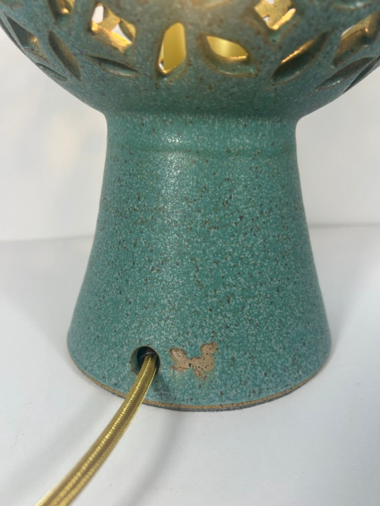 Close-up of a teal ceramic table lamp base with a gold-toned cord entering a side port, and part of a matching perforated shade visible above with warm light glowing through cutouts.