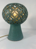 A teal ceramic table lamp with a pierced spherical ceramic shade and a tapered base, shown lit against a plain white background.