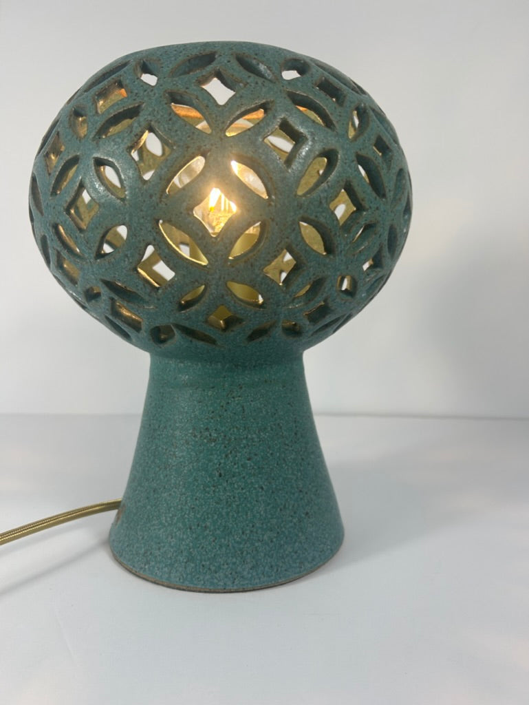 A teal ceramic table lamp with a pierced spherical ceramic shade and a tapered base, shown lit against a plain white background.