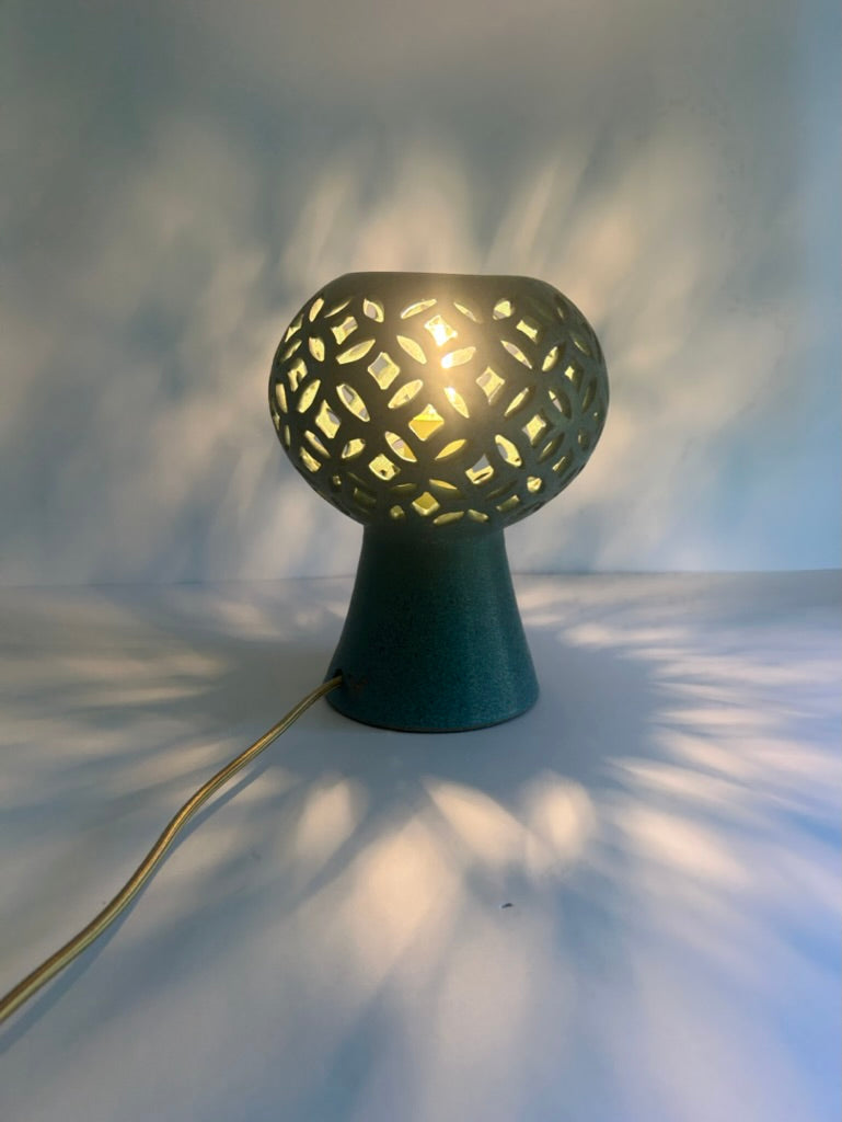 A modern ceramic table lamp with a dark teal-green base and a spherical shade featuring an intricate geometric cutout pattern that casts a star-like shadow on the wall and surface behind it. The lamp is turned on, emitting a warm yellow light, with its power cord trailing to the left.