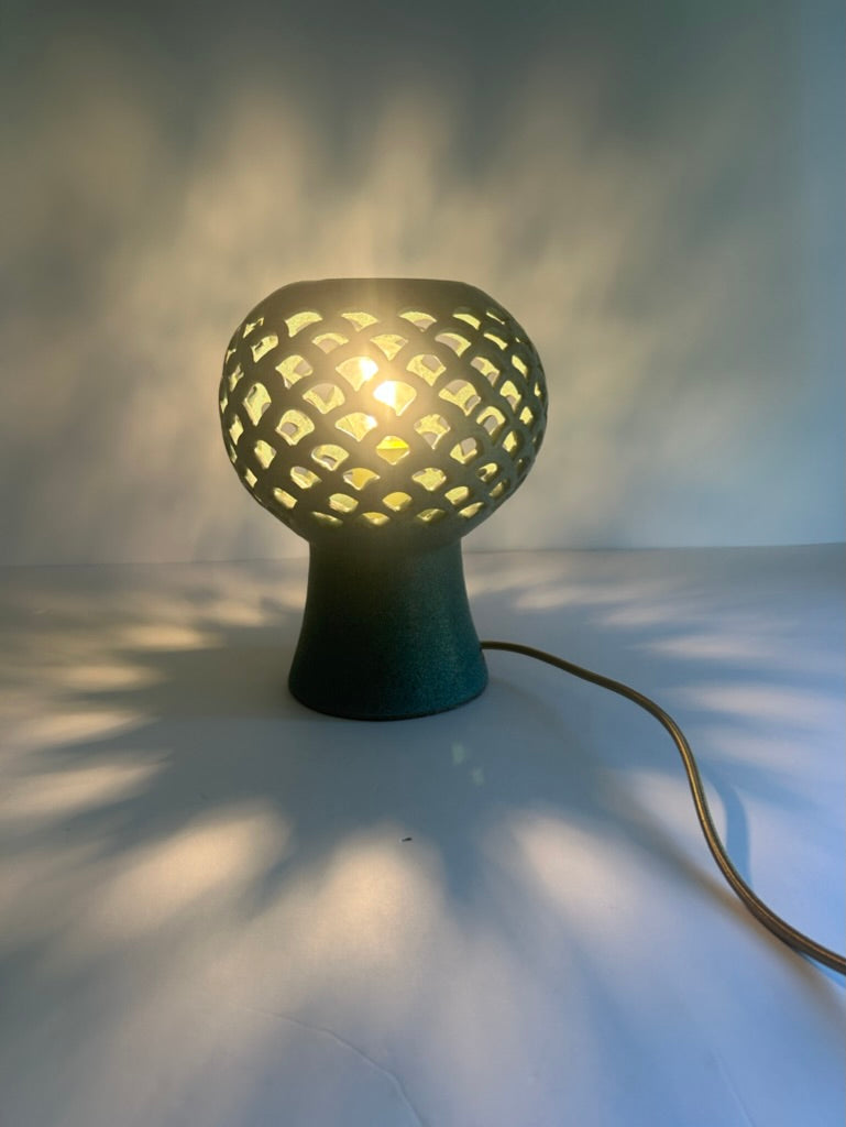 A modern ceramic  table lamp with a glowing spherical shade featuring a geometric honeycomb cutout pattern, resting on a white surface with dramatic shadow casting on a light gray background.