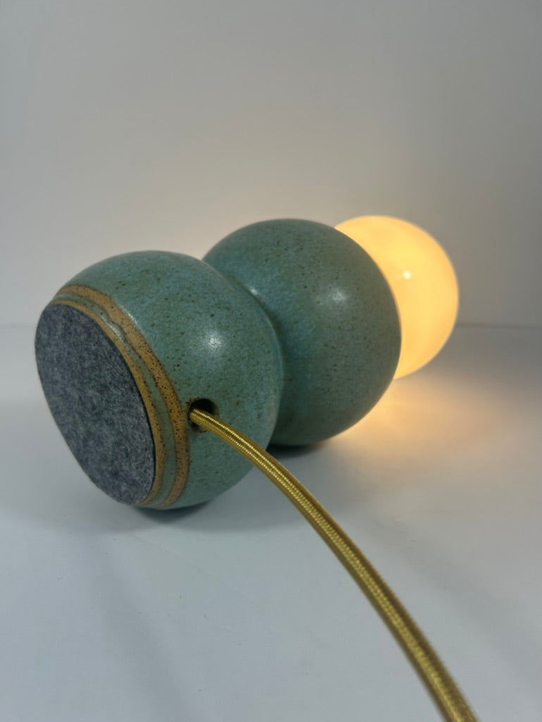 A modern two-sphere ceramic table lamp with a speckled sage green glaze, a dark gray felt end, and a warm glowing bulb inside. A gold-toned cord runs from the front sphere.