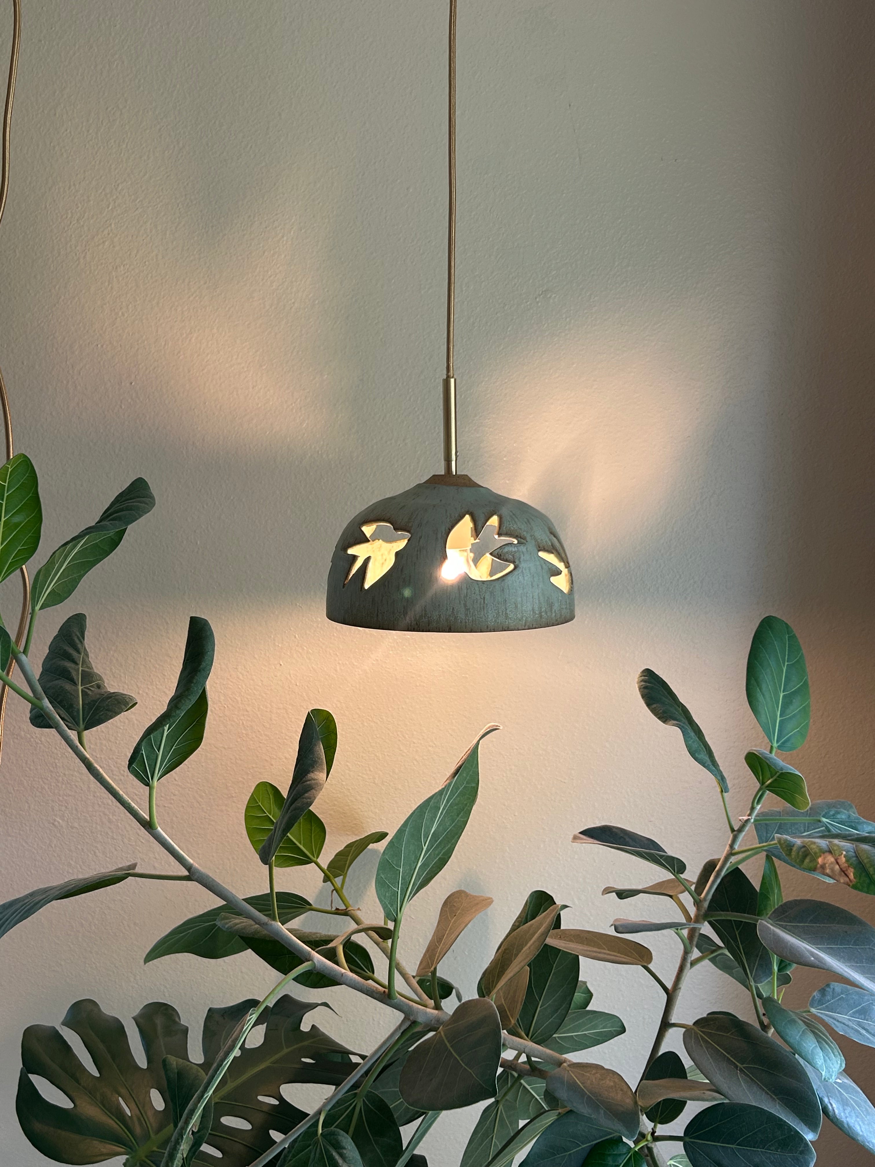 Stoneware green speckled pendant lamp with bird piercings and gold cord hanging above tree
