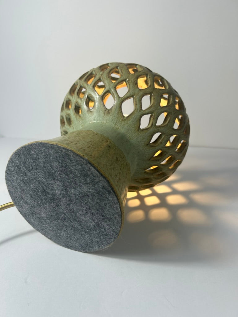 A modern sculptural table lamp with a perforated, geometric shade casting warm light, a textured sage green upper body, and a circular dark gray felt base, shown on a white surface against a white background.