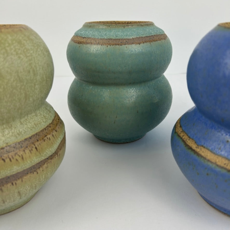 Three handmade ceramic gourd-shaped vases with textured glazes, featuring a central sage green vase and two partially visible ones in olive green and blue with brown earthy bands.